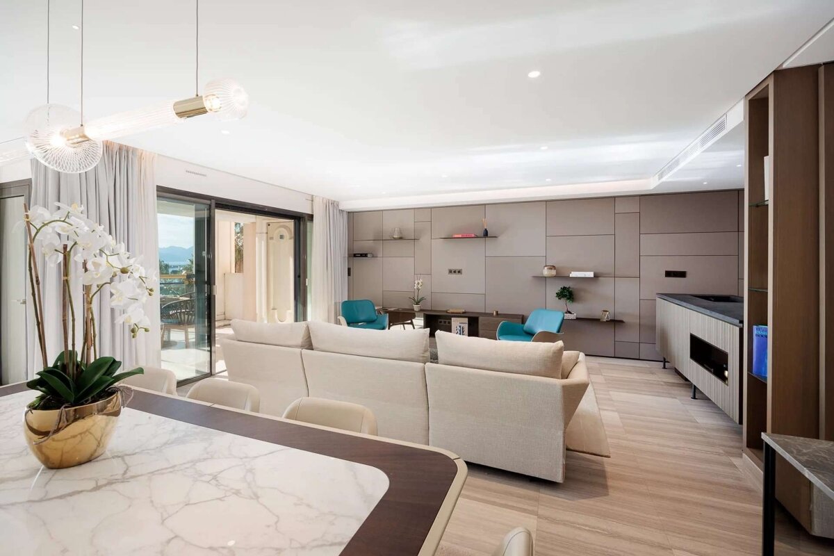 An elegant, contemporary living area with neutral decor, a large sofa, and a marble dining table. Sliding glass doors reveal a scenic outdoor view.