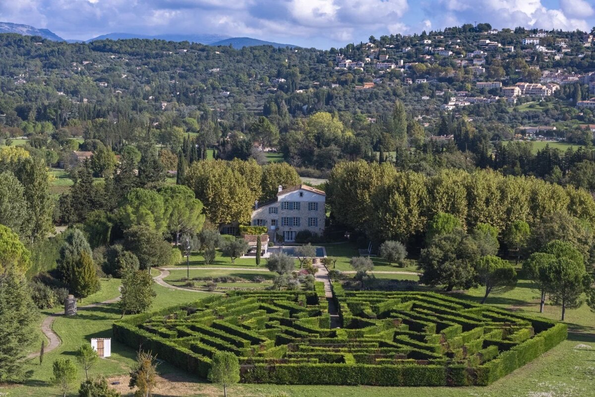 Aerial view of a grand estate featuring an intricate hedge maze, a stone house, and lush, forested hills in the background.