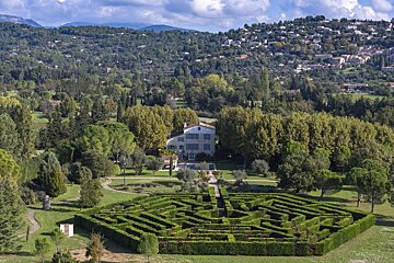 Aerial view of a grand estate featuring an intricate hedge maze, a stone house, and lush, forested hills in the background.