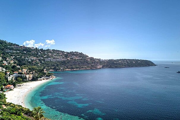 Scenic coastal view of a town built into lush green hills overlooking a vibrant blue bay with a white sand beach and distant yachts on a clear day.