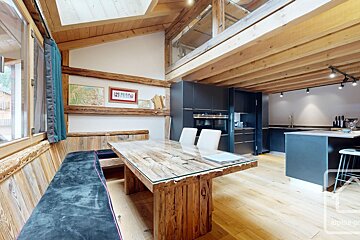A rustic-modern open-plan alpine chalet interior featuring exposed wooden beams, a large dining table, and a sleek dark kitchen.