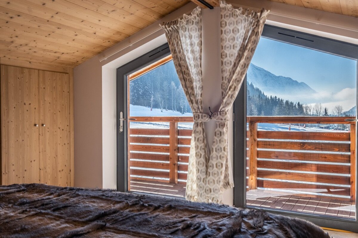 A cozy bedroom with a faux fur blanket and wooden elements, overlooking a snowy mountain landscape through a large window leading to a balcony.