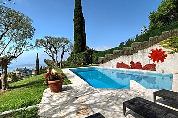 A scenic swimming pool overlooking a coastline, with lush green gardens, lounge chairs, and a painted sun wall under a clear blue sky.