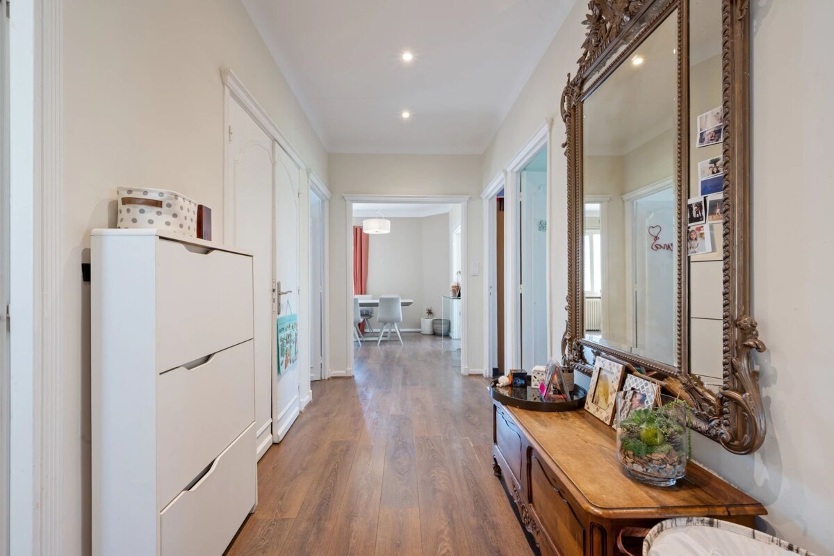 Bright hallway with wood floors, featuring a white shoe cabinet on the left and a wooden console table with an ornate mirror on the right.