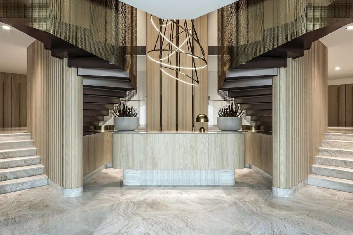 A luxurious, symmetrical interior features twin dark stairs with mesh railings, flanking a light wood reception desk. Marble floors and a modern chandelier complete the elegant design.