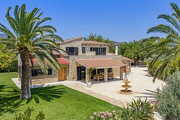 A beautiful stone villa with terracotta roofs, framed by tall palm trees, a lush green lawn, an outdoor patio, and a decorative fountain.