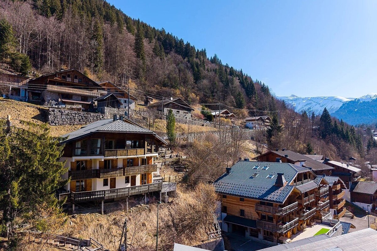 Wooden alpine chalets nestled on a hillside with pine forests, overlooking distant snow-covered mountains under a bright blue sky.