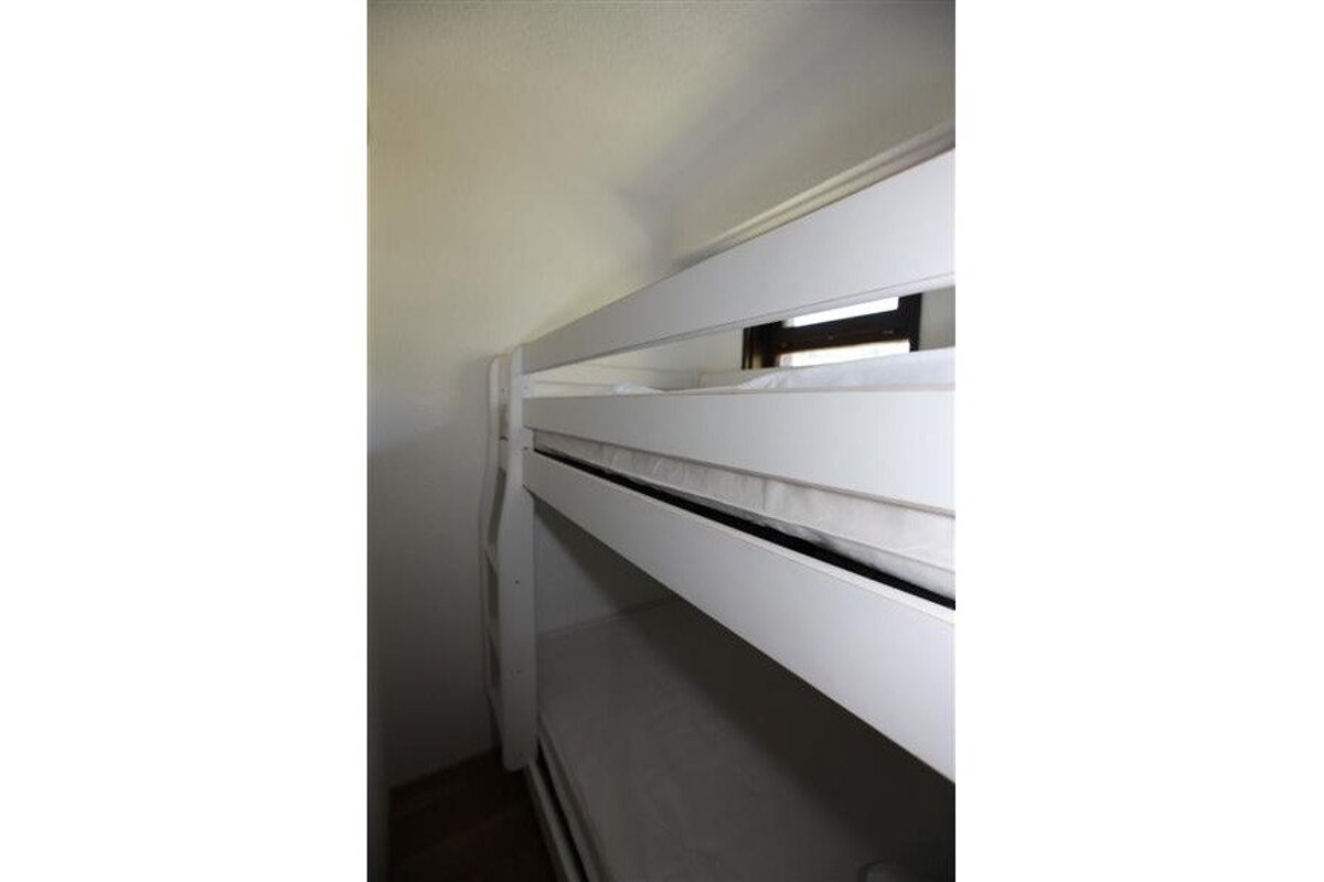 An angled view of a white bunk bed with mattresses, seen against a light-colored wall. A window is partially visible behind the top bunk.