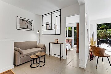 Modern open-plan apartment with light-colored tile floors, a gray couch, wooden tables, and a doorway leading to a bedroom.