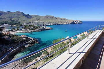Sunny panoramic view of a turquoise bay, mountains, and a resort town, framed by a modern railing.