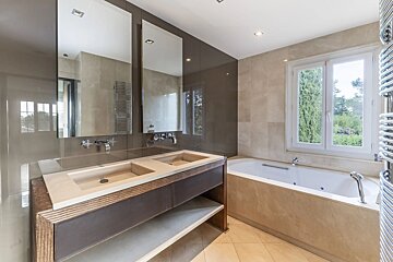 Modern bathroom with a double vanity, sleek mirrors, and a bathtub under a window with a green view, featuring light tile walls.