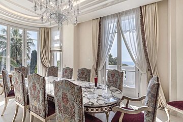 A luxurious dining room with an ornate chandelier, marble table, patterned chairs, and large windows offering views of palm trees and the sea.