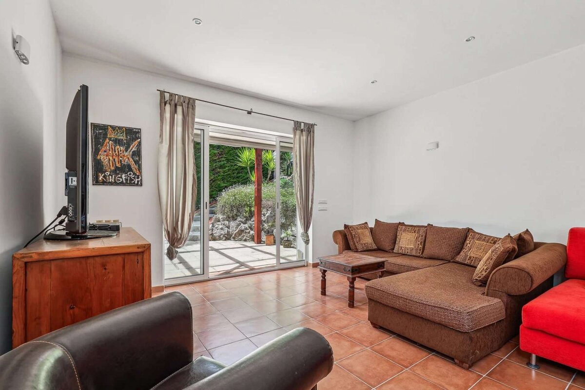 Bright living room with a brown L-shaped sofa, red chair, TV, tiled floor, and glass sliding doors revealing a lush green patio.