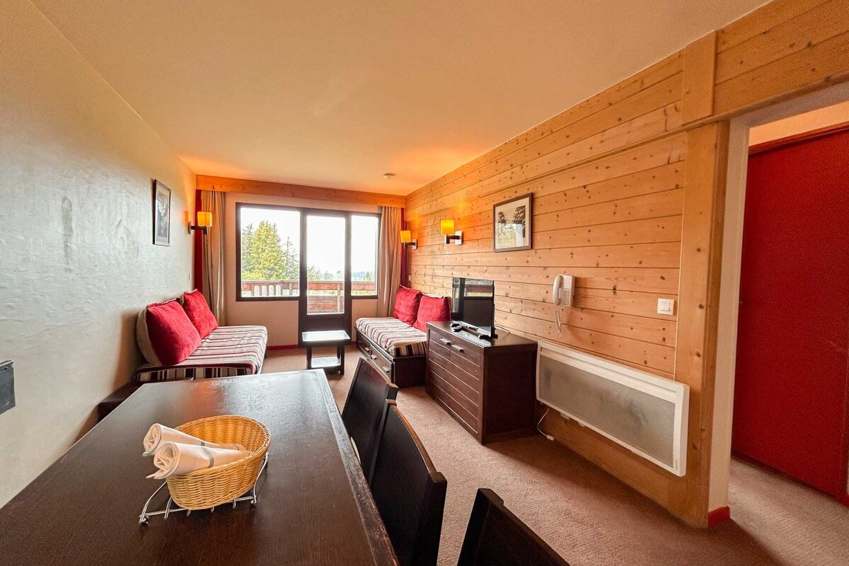 A cozy, chalet-style living area with wood paneling, two sofas, a dining table, TV, and a balcony overlooking the outdoors.