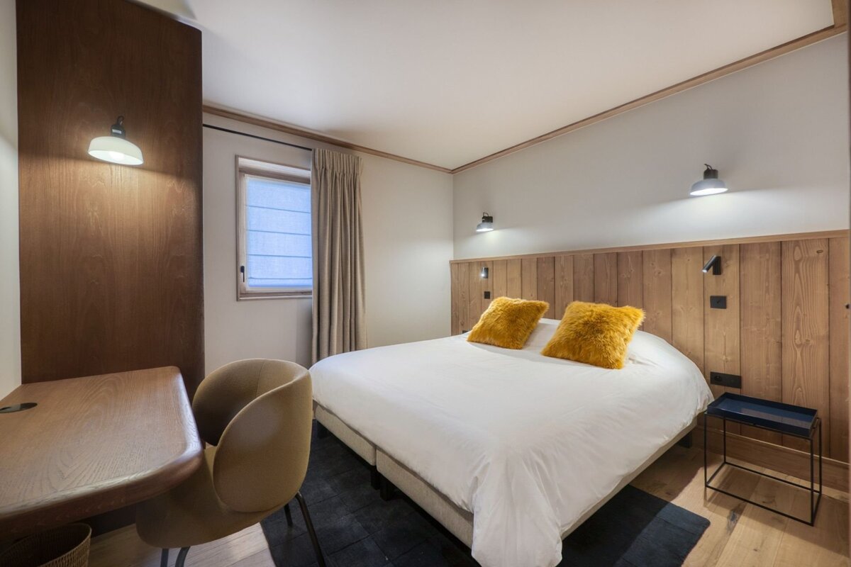 A modern, inviting bedroom with a white bed, fluffy yellow pillows, light wood paneling, a desk, and a window, illuminated by warm lighting.