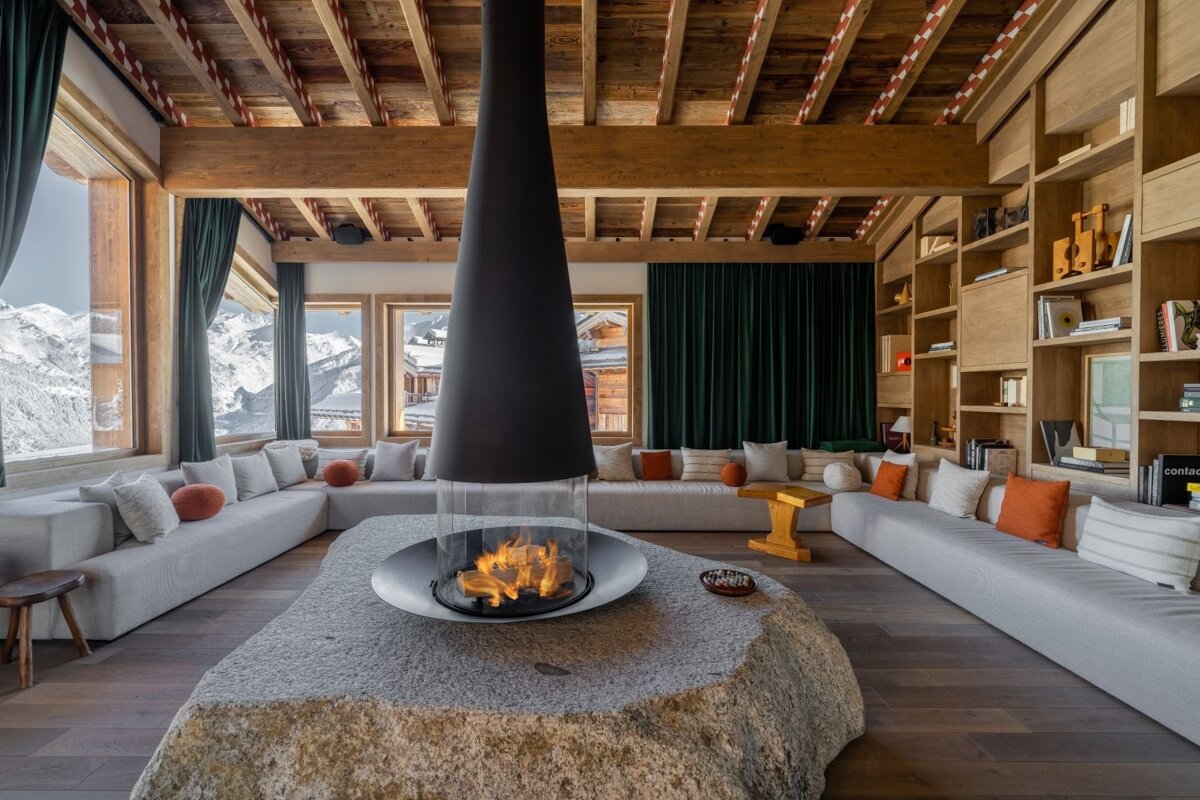 Cozy, luxurious ski chalet living room with a central fireplace, stunning snowy mountain views through large windows, and warm wooden decor.
