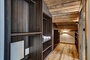 A cozy, modern rustic walk-in closet with dark wood shelving, a light wood planked ceiling and floor, illuminated by warm wall lights.