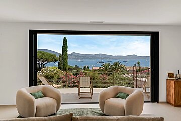 Two cozy armchairs overlook a sunny balcony and a stunning bay filled with boats, framed by a large sliding glass door.