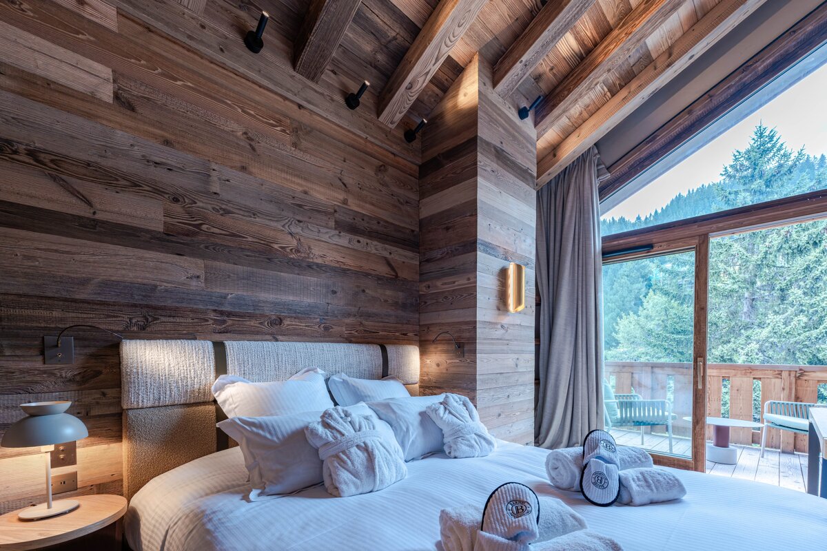A cozy, wood-paneled bedroom with white bedding, bathrobes, and slippers. A large window and balcony offer a stunning forest/mountain view.