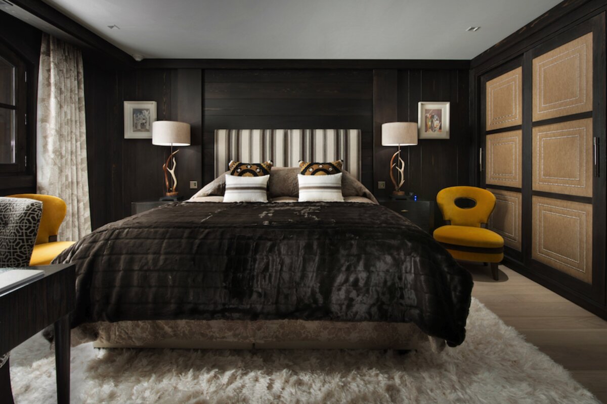 Cozy, dark wood paneled bedroom with a large bed, striped headboard, gold accent chairs, antler-like lamps, and a plush white shag rug.