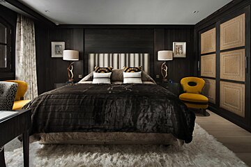 A dark, luxurious bedroom with a large bed, striped headboard, antler lamps, and bright yellow accent chairs against dark wood paneling. A white shaggy rug covers the floor.