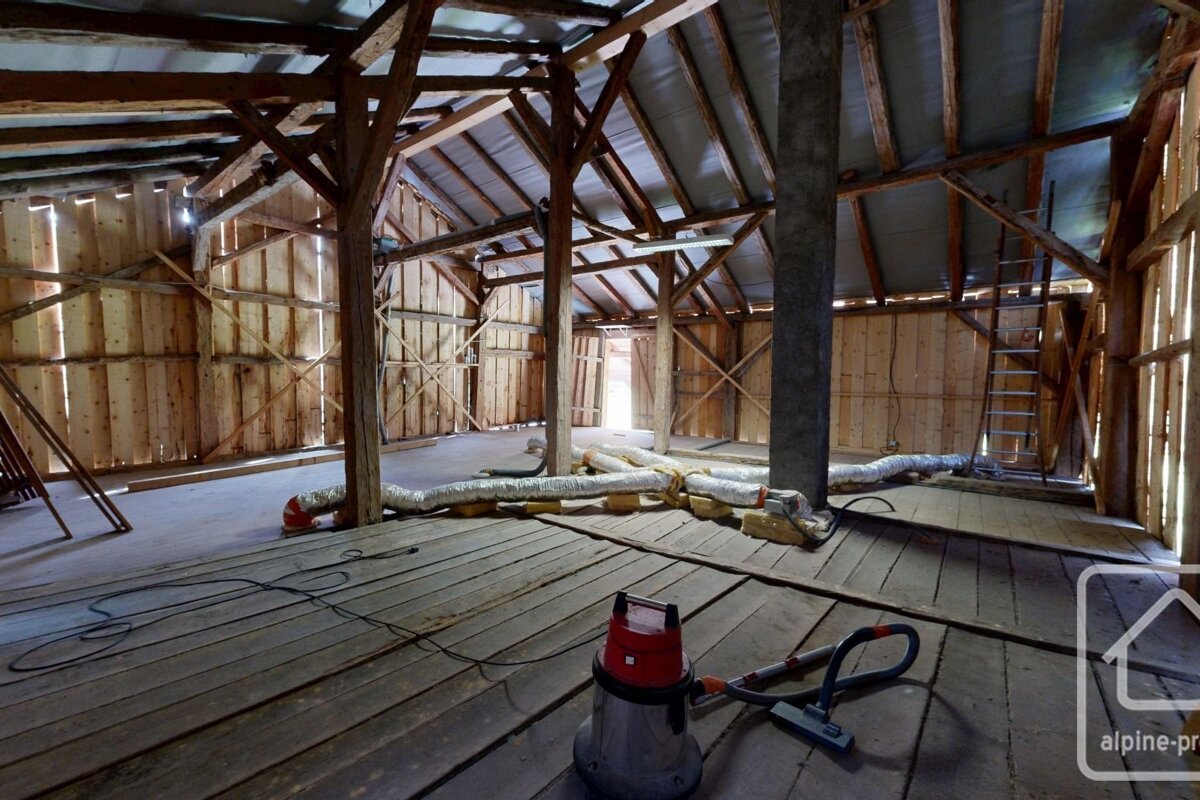Large rustic barn interior with exposed wooden beams, bare walls, a wooden floor, flexible ducts, and a vacuum cleaner. Light fills the space.