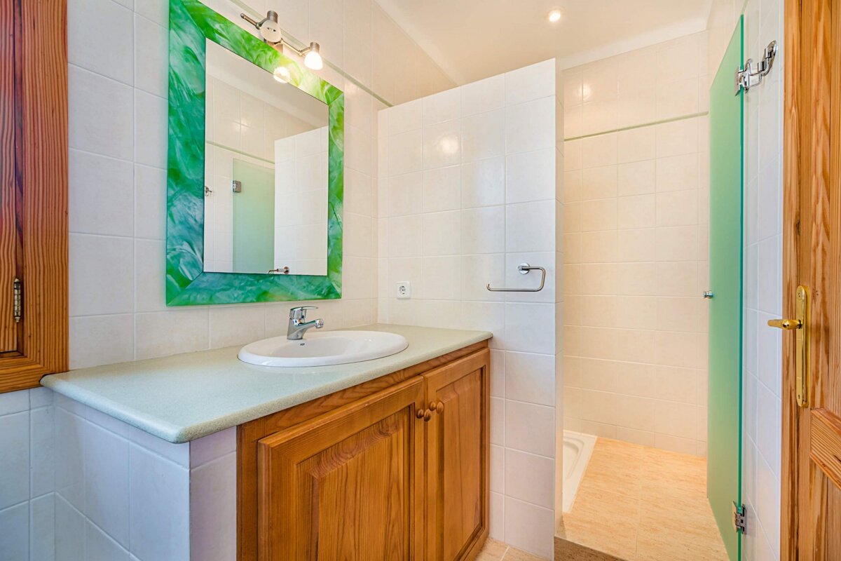 A clean bathroom with a wooden sink vanity, a striking green-framed mirror, white tiled walls, and a shower with a green glass partition.