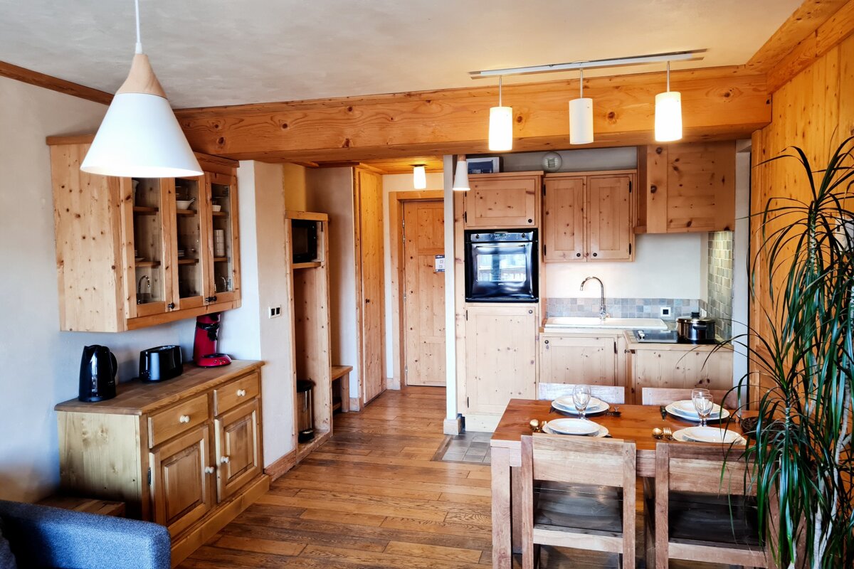 A cozy, modern rustic kitchen and dining area featuring light wood cabinets, wall panels, and flooring. A dining table is set for two.