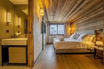 A rustic bedroom with wooden walls & ceiling, a cozy bed, and a snowy mountain view, connected to a modern bathroom with a white sink.