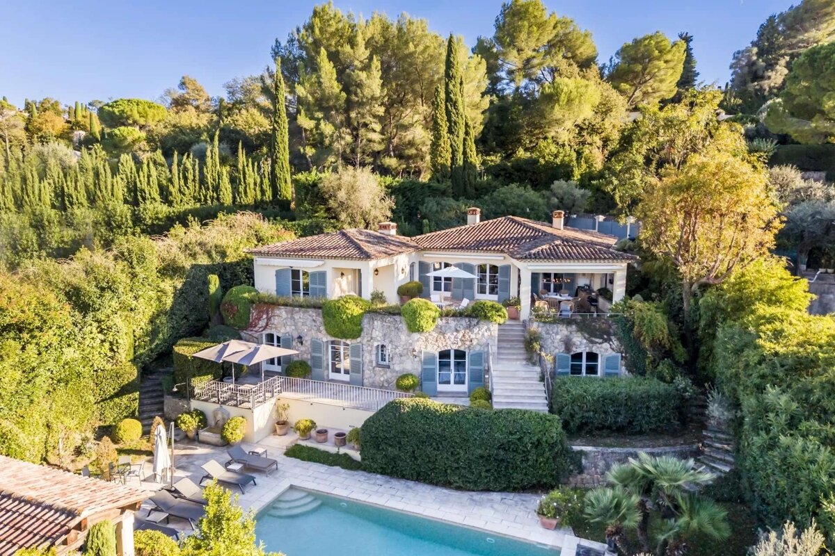 An elegant, multi-level villa with a swimming pool and terraced gardens, nestled into a lush, tree-covered hillside on a sunny day.