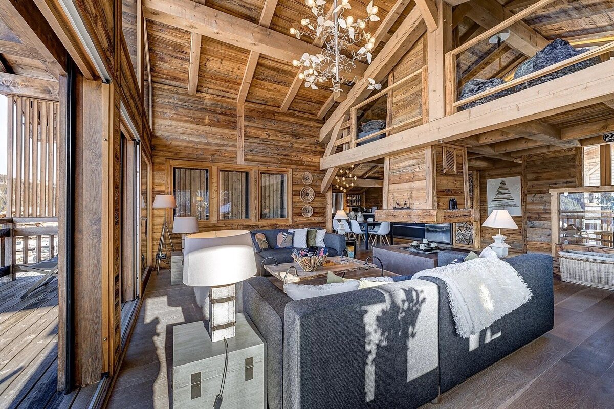 Cozy interior of a rustic wooden chalet featuring a high vaulted ceiling, modern grey sofas, a loft, and a unique chandelier.