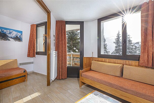 A bright room with wooden furniture and a mountain picture. Large windows reveal a sunny, snowy mountain landscape with pine trees.