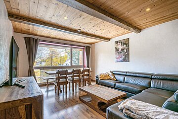 Cozy chalet living room with a leather sectional, wooden dining set, and a mountain view from the window, under a rustic wooden ceiling.