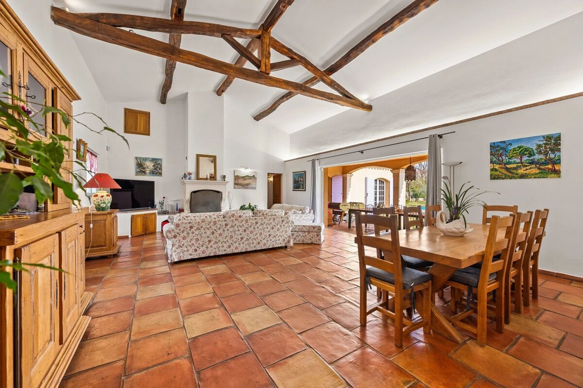 An open-plan living and dining area with terracotta tiled floors, a high ceiling with exposed wooden beams, and a dining table.