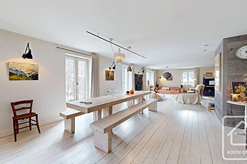 A bright, open-plan room with white wooden floors and walls. A long wooden dining table and benches dominate the foreground, with a living area beyond.