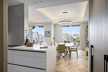 Bright modern apartment with a sleek kitchen island, dining area with green chairs, herringbone wood floors, and balcony views.