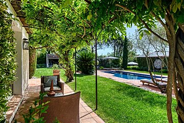 Vine-covered patio with wicker seating overlooking a green lawn and a sunny swimming pool with lounge chairs.