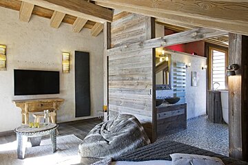 A rustic modern room with exposed wood, a TV area, cozy beanbag, and an open-plan bathroom featuring dual sinks and a tub.