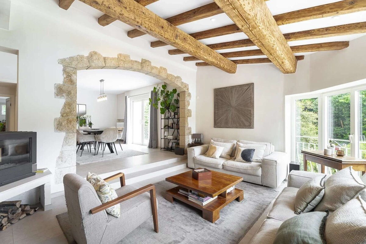 Cozy, modern rustic living room with exposed wood beams, a stone archway leading to a dining area, and bright, neutral decor.