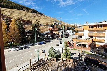 A scenic mountain village street with chalet-style buildings, cars parked along the road, and grassy hills under a blue sky.
