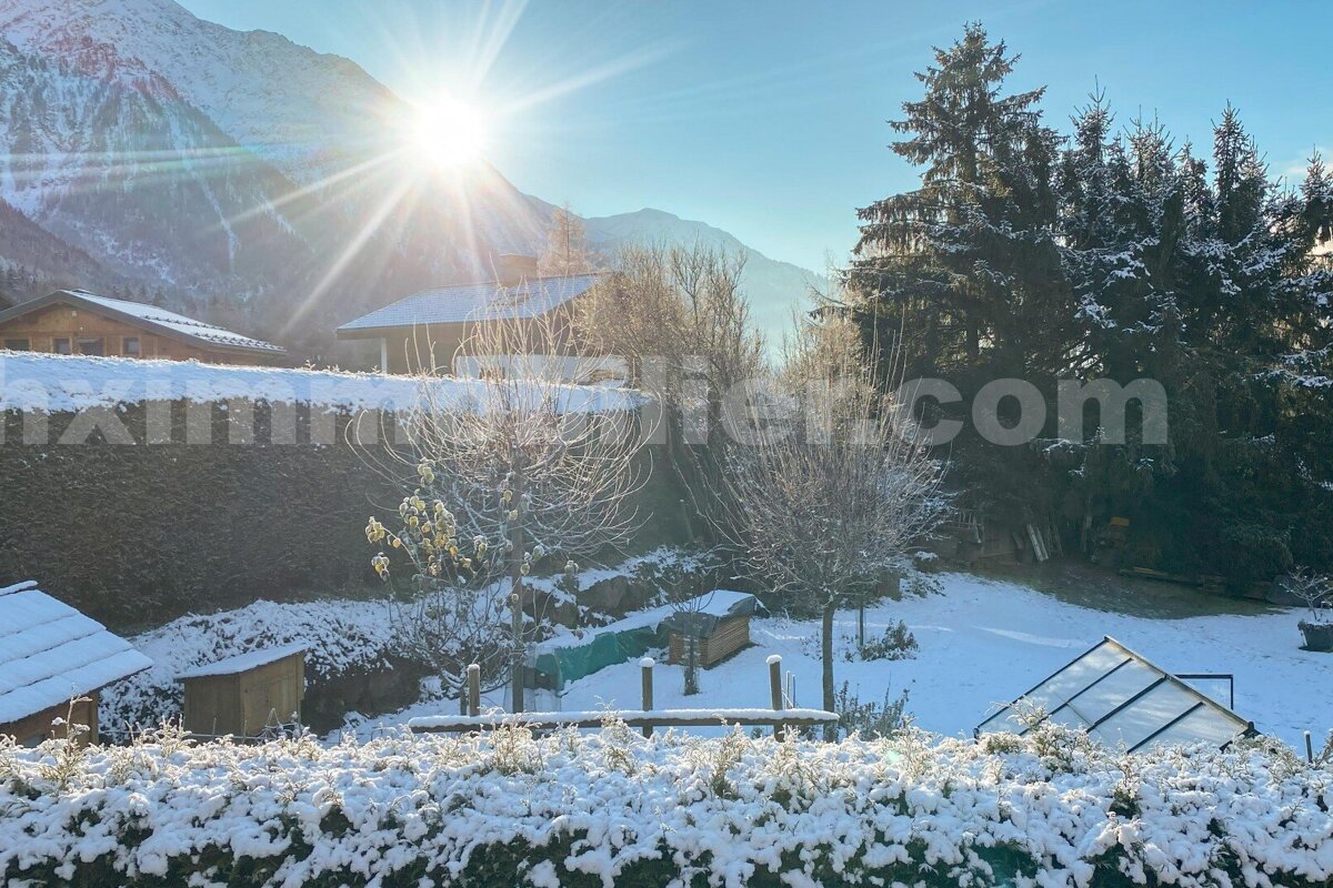 A bright, snowy mountain scene featuring sun-drenched peaks, chalets with snow-covered roofs, frosted trees, and a wintry garden.
