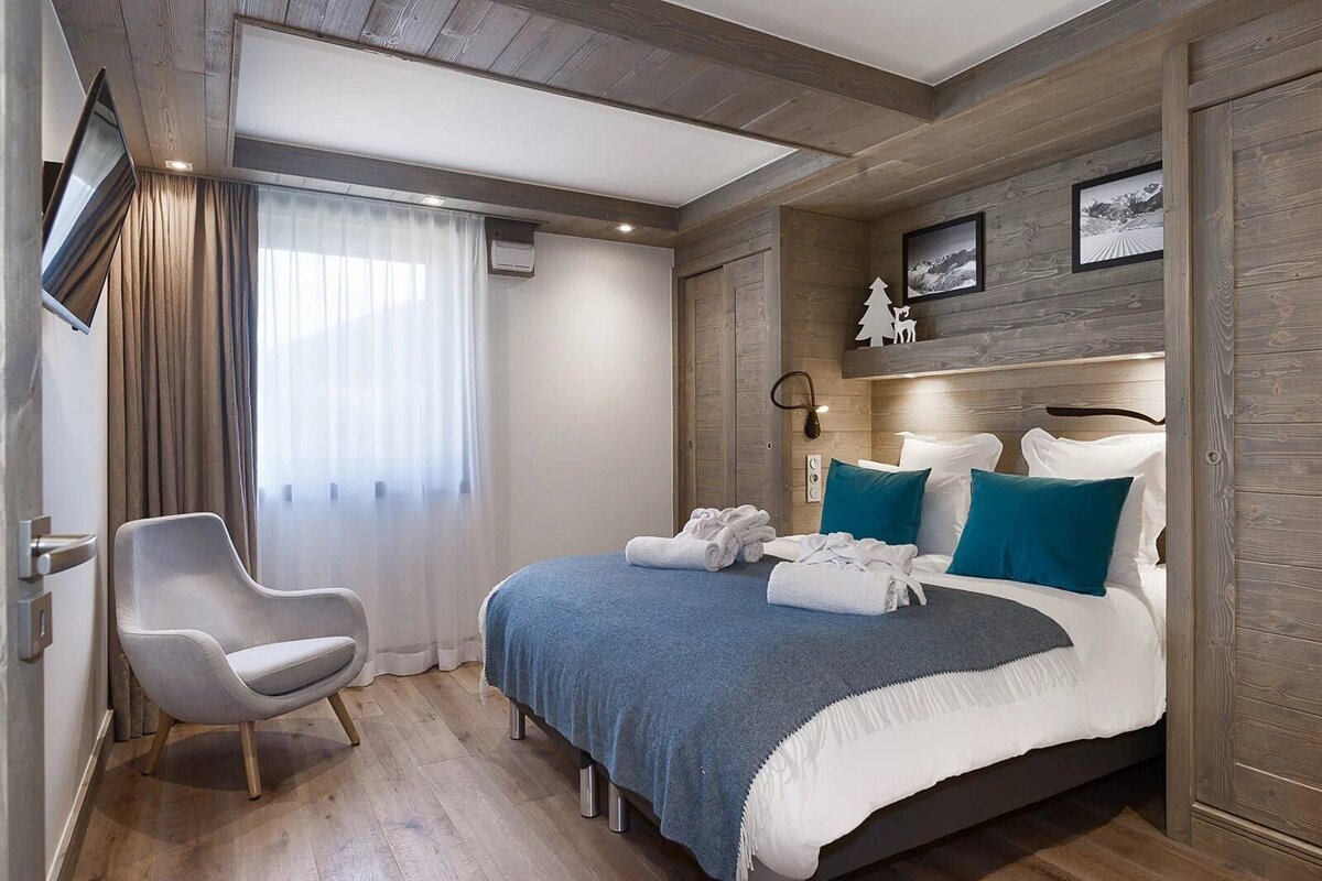 Cozy chalet bedroom with wood-paneled walls, a bed with blue pillows and throw, and a grey armchair near a window.