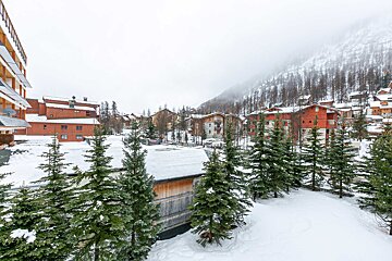 Snowy mountain village with numerous wooden buildings, evergreen trees, and a misty, forested slope under an overcast sky.