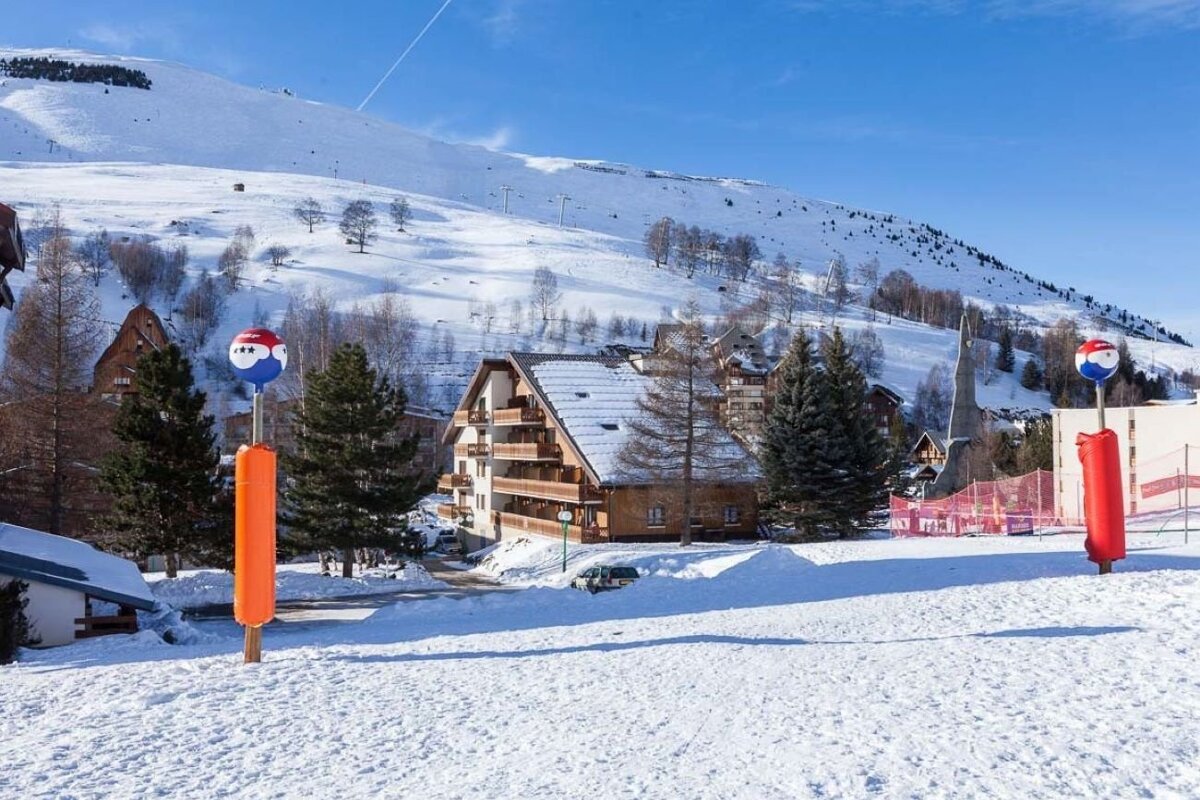 A snow-covered ski resort with wooden buildings, pine trees, and a mountain under a clear blue sky.