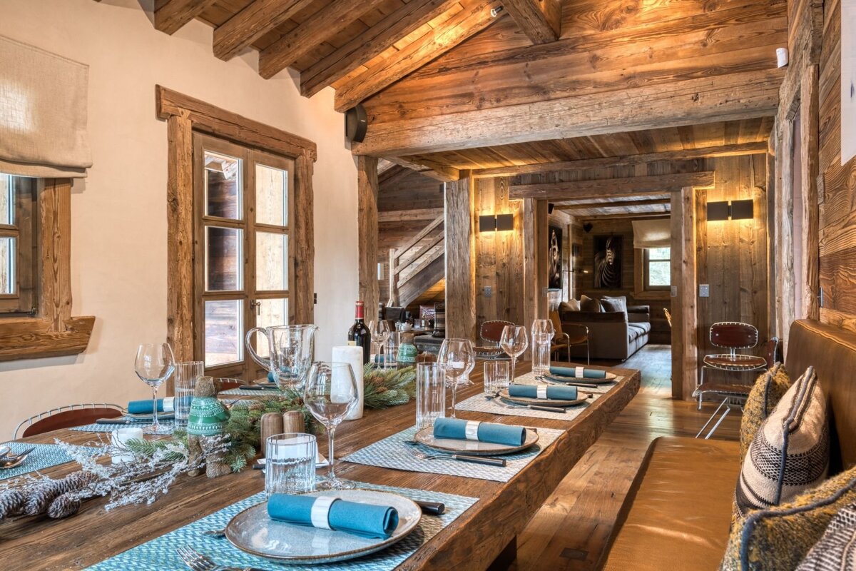 Cozy rustic chalet dining room with a long wooden table set with blue napkins. Exposed wood beams and paneling create a warm, inviting ambiance.