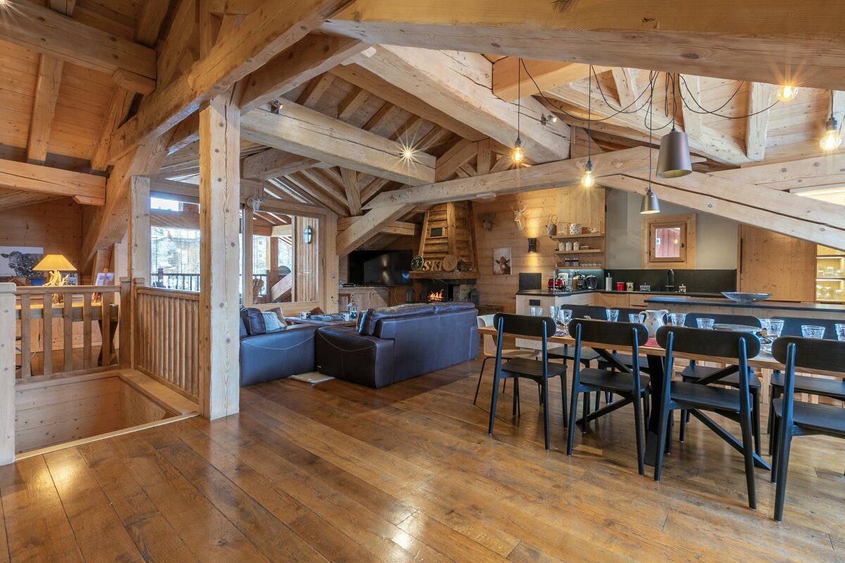 Spacious, rustic-modern wooden chalet interior with open-plan living, dining, and kitchen areas. Features exposed beams, a fireplace, and warm lighting.