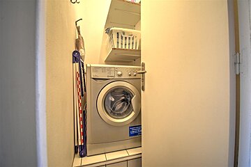 A narrow laundry closet features a front-load washing machine, a folded drying rack, shelves with a laundry basket, and a partially open door.