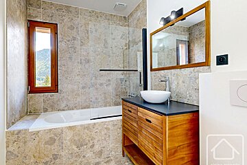 A modern bathroom features light-colored marble-look tiles, a bathtub with a mountain view window, a wooden vanity with a vessel sink, and a mirror.