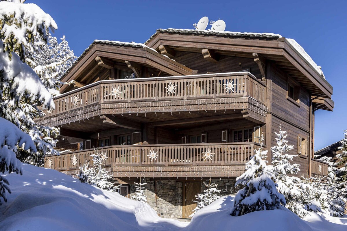 A beautiful, multi-story wooden chalet with balconies and decorative lights, surrounded by snow-covered trees under a clear blue sky.