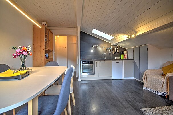 A bright attic apartment showcasing a dining area with flowers, a compact kitchenette under a skylight, and a cozy living space, all with a slanted paneled ceiling.